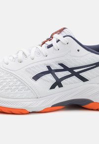 Zapatilla deportiva blanca con parte superior de malla, acentos en color marino, suela naranja y un diseño texturizado con una marca prominente en el costado.