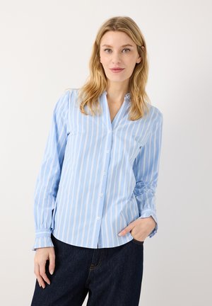 Vrouw met blond haar die een blauw-wit verticaal gestreept overhemd met knopen en donkere spijkerbroek draagt, poseert met één hand in de zak.