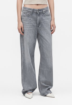 Personne portant un jean en denim gris ample, un top court beige et blanc rayé, et des chaussures blanches pointues sur un fond uni.