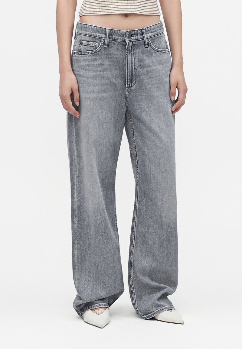 Personne portant un jean en denim gris ample, un top court beige et blanc rayé, et des chaussures blanches pointues sur un fond uni.