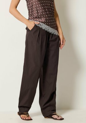 Person trägt weite dunkelbraune Hose, braunes ärmelloses Oberteil mit Spitzenbesatz und Polka-Dots sowie braune Sandalen und steht mit den Händen in den Taschen.