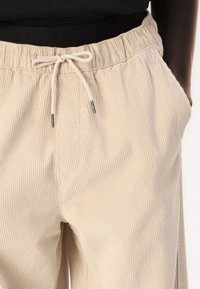 Beige corduroy pants with an elastic waistband and drawstring, featuring vertical ribbed texture and side pockets.