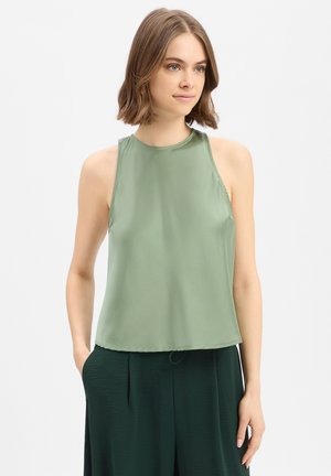 Jonge vrouw met bruin schouderlang haar, die een mouwloos lichtgroen topje en donkergroene broek draagt, staand met handen in de zakken.