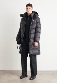 Schwarze Daunenjacke mit langer Passform, Reißverschluss und Kapuze. Getragen über einem schwarzen Hemd mit Knöpfen und Hose, dazu robuste schwarze Schuhe.