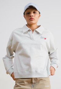Svetlož grey oversized mikina s golierom, ktorá má malé červené logo. Model má na sebe svetlomodrú čiapku a béžové nohavice s brandingom.