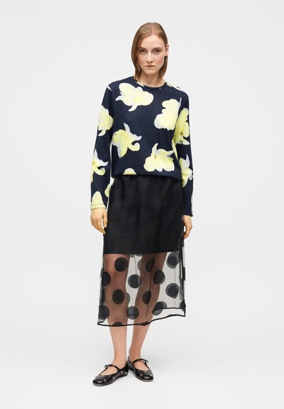 Γαλάζιο πουλόβερ με κίτρινα λουλουδάτα σχέδια, φορεμένο πάνω από μαύρη διάφανη φούστα με πουά. Μαύρα φλατ παπούτσια ολοκληρώνουν το σύνολο.