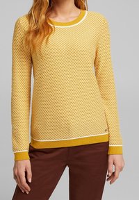 Gelber Strickpullover mit strukturiertem Muster, weißen Akzenten am Halsauschnitt und Saum sowie langen Ärmeln. Kombiniert mit braunen Hosen.