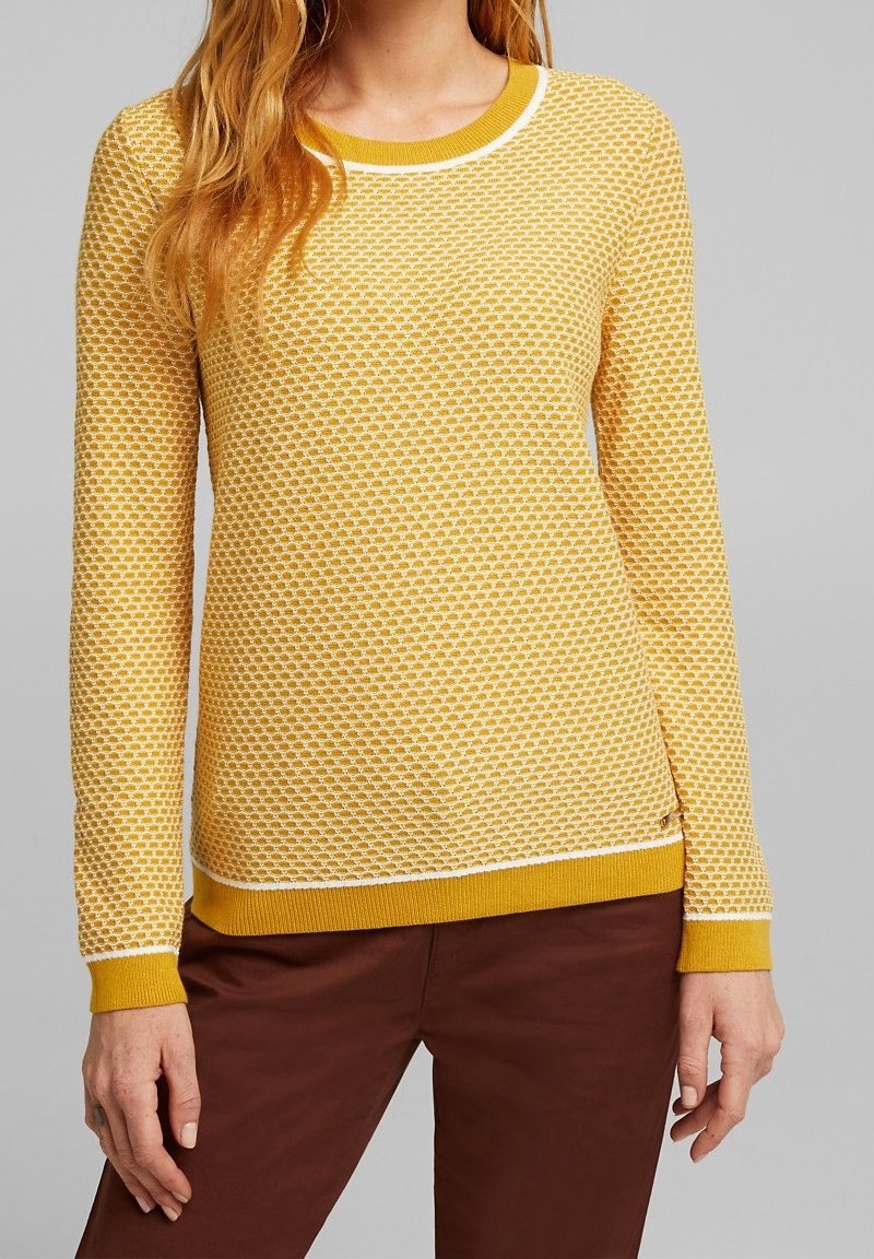 Gelber Strickpullover mit strukturiertem Muster, weißen Akzenten am Halsauschnitt und Saum sowie langen Ärmeln. Kombiniert mit braunen Hosen.