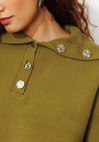 Sweat à capuche vert olive, avec un tissu texturé, quatre grands boutons métalliques et un détail de col.