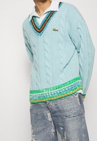 Ljusblå stickad tröja med V-ringning, kabelmönster och en flerfärgad randig nederkant. Bärs över en skjorta med krage.