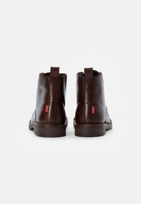 Un par de botas de tobillo de cuero marrón oscuro con tiradores y etiquetas del logo rojo de Levi's en la parte posterior, presentadas sobre un fondo blanco.