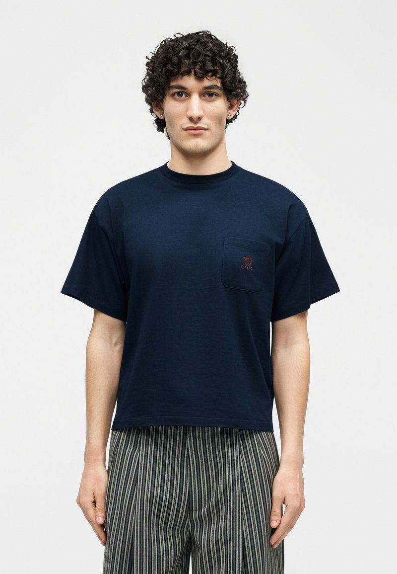 Giovane uomo con capelli ricci che indossa una maglietta blu navy con taschino sul petto e pantaloni a righe verticali bianche e nere, in piedi davanti a uno sfondo semplice.