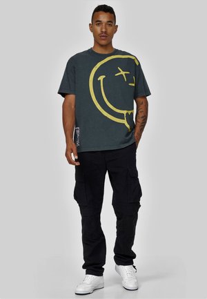 Mørkegrå kortærmet t-shirt med stort gult smilende ansigt grafik, parret med løse sorte cargo bukser og hvide sneakers.