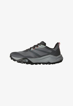 Zapatilla de trail running en gris y negro con suela resistente, cordones delanteros, lengüeta roja para tirar y logo de The North Face en el lateral de la suela.