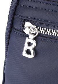 Tessuto blu navy con finitura testurizzata, caratterizzato da un tirante della cerniera argentato a forma della lettera "B". Primo piano del dettaglio della cerniera.