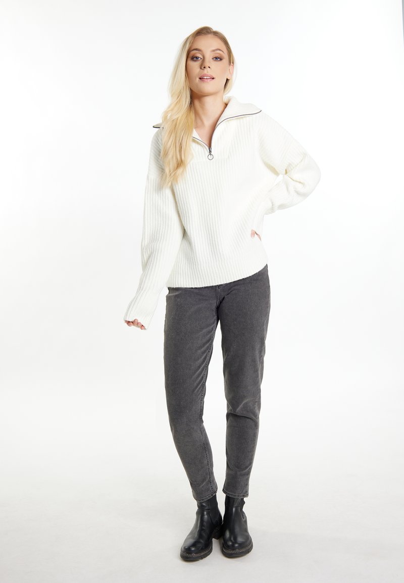 Weißer gerippter Pullover mit einem Reißverschlusskragen, kombiniert mit grauen Hosen und schwarzen Knöchelstiefeln. Glatte Textur und lässiger Schnitt.