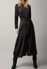Robe à manches longues, de couleur sombre avec un motif léopard, taille ceinturée et jupe fluide. Portée avec des bottes noires jusqu'aux genoux, avec un col.