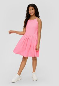 Rosa ärmelloses Kleid aus strukturiertem Stoff, mit ausgestellt Rock und geraffter Taille. Kombiniert mit weißen Sneakers vor einem neutralen Hintergrund.