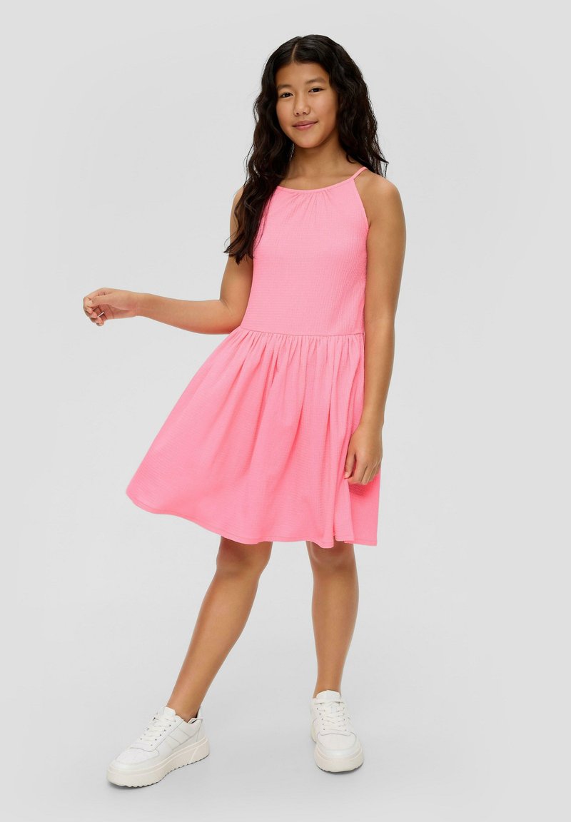 Rosa ärmelloses Kleid aus strukturiertem Stoff, mit ausgestellt Rock und geraffter Taille. Kombiniert mit weißen Sneakers vor einem neutralen Hintergrund.