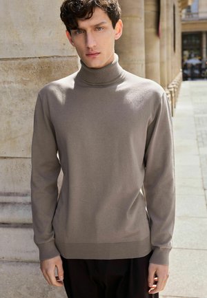 Pull en col roulé beige en tricot doux et lisse avec des manches longues, des poignets et un ourlet côtelés, porté avec un pantalon sombre devant un fond en pierre.