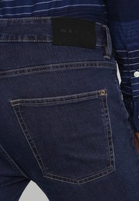 Tmavě modré džíny z denimu mají obdélníkovou zadní kapsu, kontrastní švy a černou koženou nášivku s nápisem "WĀVĒ."