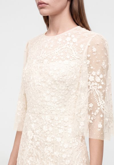 Femme portant une robe en dentelle blanche transparente avec une broderie florale complexe et des manches trois-quarts sur un fond uni.