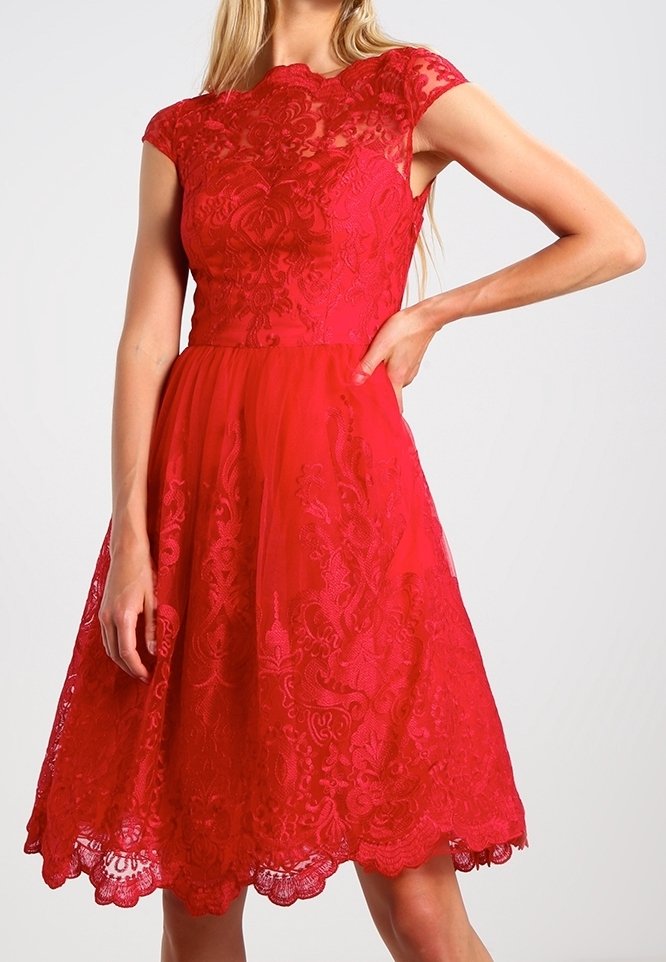 Robe rouge en dentelle avec des manches courtes, un corsage ajusté et une jupe évasée. L'ourlet présente un bord festonné et des détails floraux élaborés.