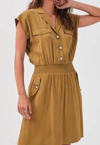 Robe courte en khaki à manches courtes avec un devant à boutons, taille smockée et poches plaquées. Fabriquée en tissu doux, elle possède un col.
