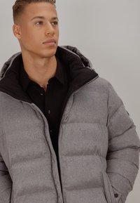 Grå dunjakke med sort hette, teksturert stoff, glidelåslukking og store polstrede seksjoner. Brukt over en svart skjorte med knapper.