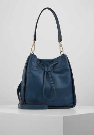 Sac seau en cuir bleu à texture, avec une bandoulière réglable, une fermeture par cordon de serrage et des fermoirs en métal doré, posé sur une surface blanche.