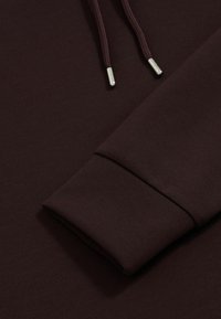 Bordowa bluza z kapturem o miękkiej teksturze, z włóknami ściągaczy oraz sznurkami z metalowymi końcówkami. Prosty design, bez widocznych wzorów.
