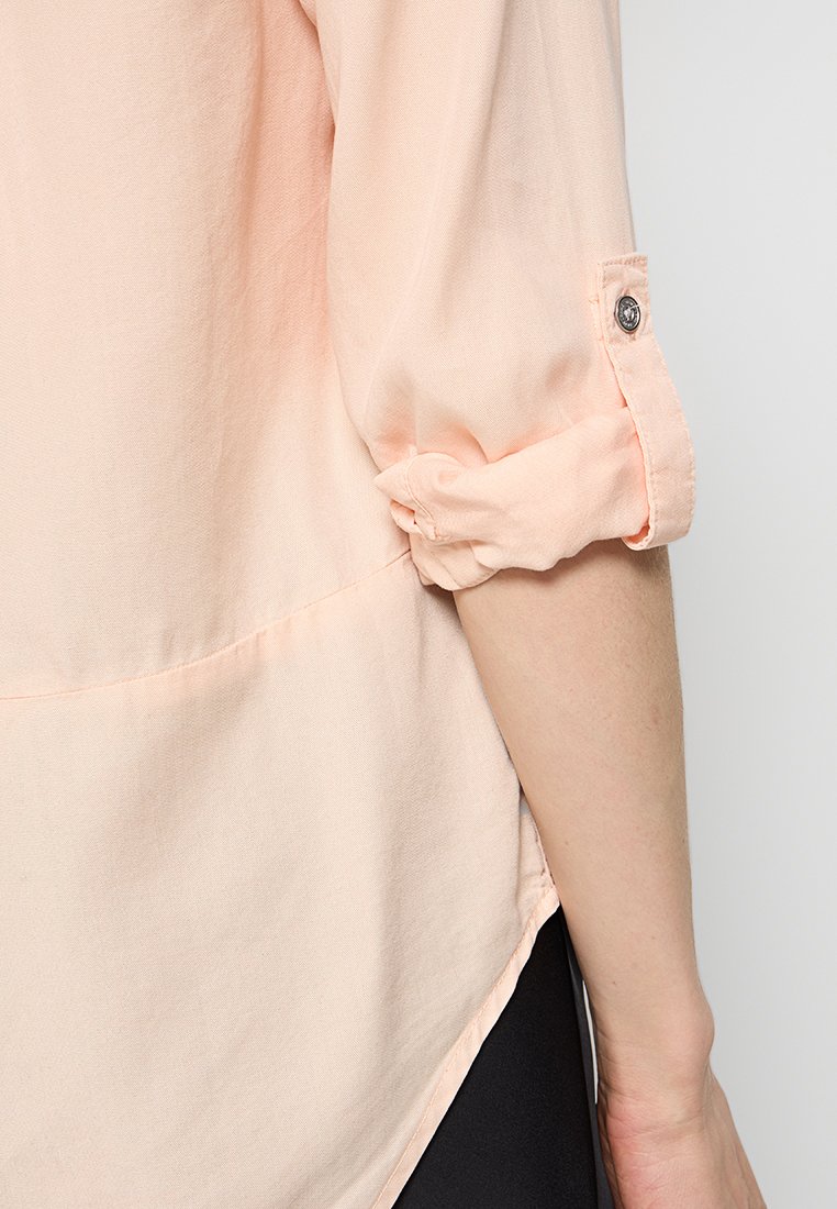 Blusa abotonada de color melocotón claro con mangas enrolladas, confeccionada en una suave tela texturizada. Presenta un dobladillo curvado y un sutil detalle de costura.