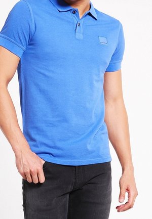 Mann trägt ein blaues Poloshirt mit kurzen Ärmeln und schwarze Jeans, steht vor einem weißen Hintergrund.
