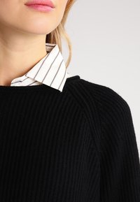 Schwarzer gerippter Pullover mit weitem Ausschnitt, über einem weißen gestreiften Hemd mit Kragen getragen, das strukturierten Stoff und kontrastierende Muster zeigt.
