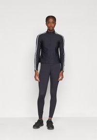 Giacca sportiva nera con righe bianche laterali, zip completa, abbinata a leggings scuri e scarpe nere. Tessuto liscio e aderente, maniche lunghe.
