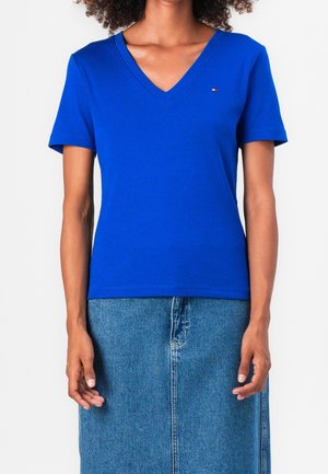 Femme portant un t-shirt bleu vif à manches courtes avec un col en V et une jupe en jean bleue mi-longue, debout avec les bras détendus.