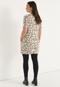 Kurzärmliges Kleid in Hellbraun mit einem weißen abstrakten Muster. Kombiniert mit schwarzen Leggings und Stiefeletten, zeigt seitliche Taschen.
