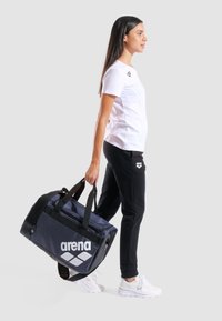 Bolso de lona azul oscuro con gran logo blanco de "arena", asas resistentes, lados de malla y cierre de cremallera. El modelo lleva una camiseta blanca y pantalones negros.