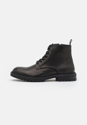 Bota negra de cuero para el tobillo con cordones en la parte delantera, lengüeta en el talón y suela gruesa con relieve sobre fondo blanco.