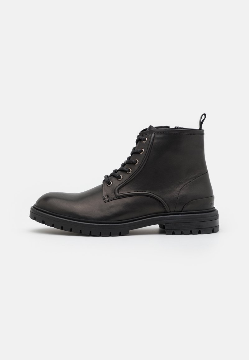 Bottine en cuir noir avec lacets à l'avant, languette au talon, et semelle épaisse crantée sur fond blanc.