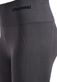 Close-up van donkergrijze naadloze sportleggings met getextureerde geribbelde patronen op de dij en een brede tailleband met zwart "hummel" logo.