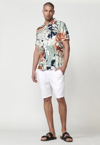 Camisa de manga corta con estampado tropical multicolor, combinada con pantalones cortos blancos. El modelo lleva sandalias negras. El fondo es neutro.