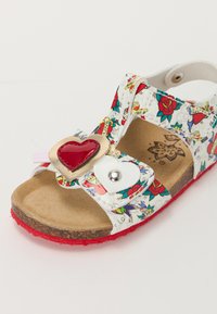 White sandal with colorful floral pattern, red heart accent, velcro straps, and textured brown insole. Red outsole for added grip.