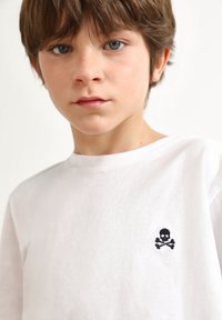 Camiseta de algodón blanca con cuello redondo, que presenta un bordado de una calavera y huesos cruzados en la parte izquierda del pecho.
