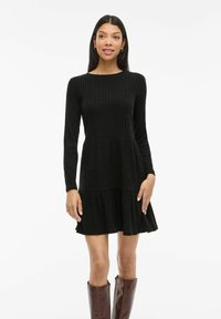 Femme portant une robe longue à manches ribbed noire avec une jupe évasée et des bottes marron jusqu'aux genoux, se tenant devant un fond blanc uni.