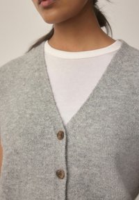 Gros plan sur une personne portant un t-shirt blanc à col rond sous un gilet tricoté gris clair à boutons avec des boutons marron.