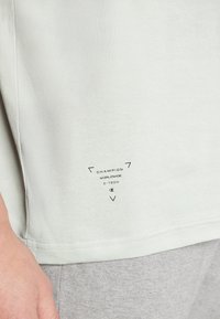 Světlé šedé tričko s krátkým rukávem a hladkým povrchem, se černým potiskem značky "Champion Worldwide C-Tech" poblíž lemu.