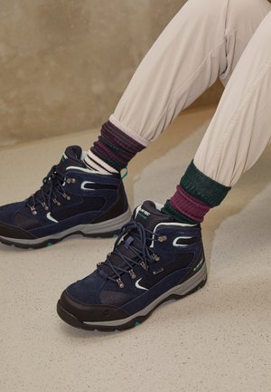 Personne portant des bottes de randonnée bleu marine à semelles noires, des chaussettes rayées multicolores et un pantalon beige clair, assise sur un sol clair.