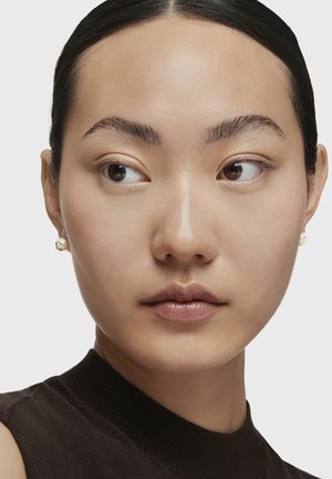 Orecchini a perno in oro e perla mostrano una forma arrotondata con una finitura testurizzata, indossati con un top nero senza maniche su uno sfondo chiaro.