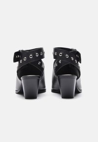 Pari mustia nahka korkokenkiä leveillä nilkkavöillä, joissa on hopeiset niitit, kuvattuna takaa valkoista taustaa vasten.